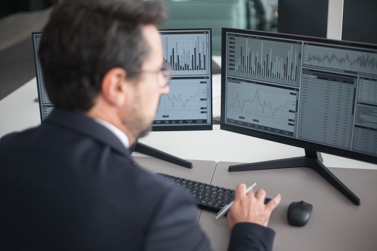 A businessman in a suit analyzing financial charts and graphs on two computer monitors indoors.